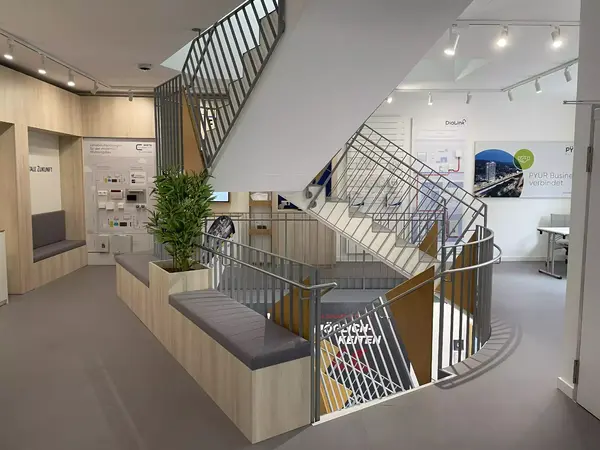Modernes Büro mit zentraler Treppe, Sitzbereich, Infotafeln und Pflanzen im Eingangsbereich.